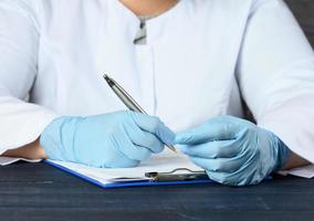 a doctor in a white coat and blue medical gloves sits at the table and writes with a pen photo