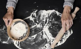 Female hands hold a round wooden sieve with white wheat flour on a black table, top view photo