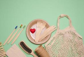 white woven cotton bag, paper cups and wooden toothbrushes on a green background. Recyclable waste, top view photo