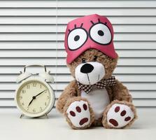 brown teddy bear sits with a bandage for sleeping on a white table. Near the alarm clock, time is seven in the morning, difficulty getting up in the morning photo