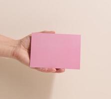 Female hand holding empty pink paper on a beige background. Copy paste image or text, close up photo