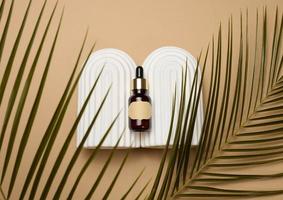 glass brown bottle with pipette and paper blank label on light brown background, near palm leaf. Mockup skincare photo