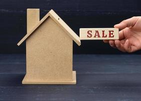 wooden model of a house and a hand holds a block with an inscription  sale on a blue background. House sale concept, real estate investment photo