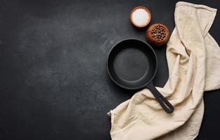 Empty black round cast iron frying pan with handle on black table, top view. Copy space photo