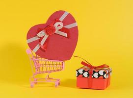 heart shaped cardboard gift box in miniature metal trolley on yellow background. Festive backdrop, photo