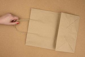 female hand holds an empty brown paper bag by the handles on a brown background, rejection of plastic bags photo