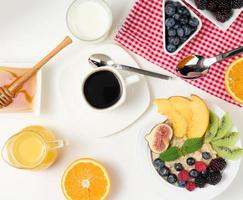 taza de café negro, un plato de avena y fruta, miel y un vaso de leche en una mesa blanca, desayuno saludable por la mañana, vista superior foto