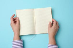 two female hands hold an open notebook with blank white sheets on a blue background, top view photo