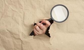 a woman's hand holds a glass magnifier, part of the body sticks out of a hole in brown paper photo