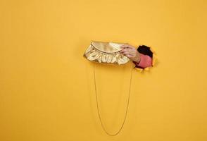 female hand is holding a small golden clutch bag with a metal chain on a yellow background. Part of the body sticking out of a torn hole in a paper background photo