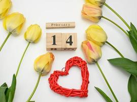 tulipanes amarillos, calendario de madera con fecha 14 de febrero y corazón rojo sobre fondo blanco foto