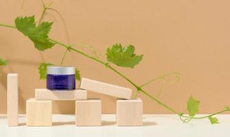 cosmetic products in a blue glass jar with a gray lid on a wooden podium made of cubes, behind a branch of grapes with green leaves. Blank for branding products, moisturizer on beige background photo