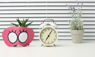 pink textile mask and round white alarm clock on the table, behind the white blinds photo