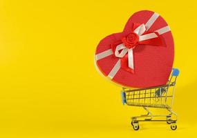 heart shaped cardboard gift box in miniature metal trolley on yellow background. Festive backdrop photo