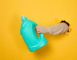 blue plastic bottle with liquid detergent in a female hand on a yellow background. A part of the body sticks out of a torn hole in the background photo