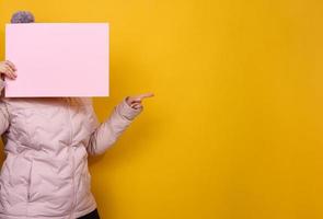 woman in pink winter jacket and hat holds blank pink sheet of paper on yellow background. Seasonal sale. Place for inscription photo