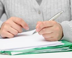 una mujer de apariencia caucásica se sienta con ropa gris en una mesa blanca y firma documentos con un bolígrafo de metal. empresario en el trabajo, firma en contrato y factura foto