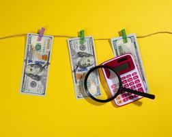 one hundred US dollar bills hang on a rope with clothespins, magnifier and calculator, yellow background. Money laundering concept photo