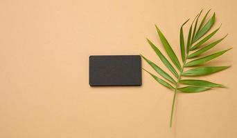 a stack of rectangular black paper business cards and a leaf of a palm tree on a brown background. View from above photo