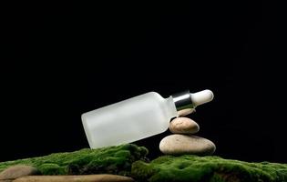 botella de vidrio blanco con soportes de pipeta sobre musgo verde, fondo negro. marca de spa de cosméticos. embalaje para gel, suero, publicidad y promoción de productos, maqueta foto
