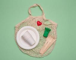 white cotton bag, paper cups and wooden forks and spoons on a green background. Recyclable waste, top view photo