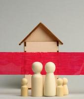 familia de figuras de madera y una casa de madera detrás de una cinta roja sobre un fondo gris. concepto de cuarentena foto