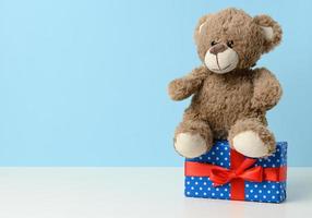 cute brown teddy bear holding a box wrapped in blue paper and red silk ribbon on white table. Prize and congratulations photo