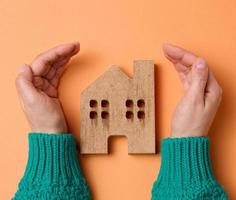 manos femeninas dobladas entre sí sobre una casa modelo en miniatura de madera sobre un fondo marrón. concepto de seguro inmobiliario, protección del medio ambiente, felicidad familiar foto