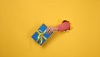 female hand holds a box with a gift on a yellow background, part of the body sticks out of a torn hole in a paper background. Congratulation, holiday surprise photo