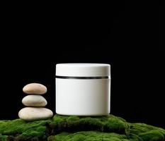 round white plastic jar with lid for cosmetics stands on green moss, black background. Natural creams and masks. Product branding photo