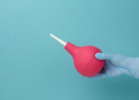 a hand in a blue medical glove holds a pink rubber enema on a blue background photo