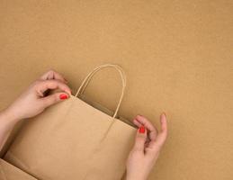 female hand holds an empty brown paper bag by the handles on a brown background photo