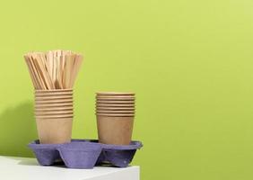 stack of brown disposable paper cups and a stand on a white table, green background. Utensils for takeaway drinks, zero waste photo