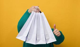 a woman in a sweater holds white paper bags on a yellow background and shows a like gesture with her hand. Purchases. Seasonal sale photo
