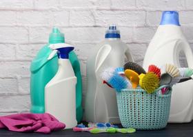 esponjas, cepillos de plástico y botellas de detergentes sobre una mesa de madera azul. artículos de limpieza del hogar sobre fondo de pared de ladrillo blanco foto