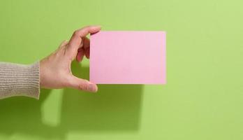 Female hand holding empty pink paper on a green background. Copy paste image or text, close up photo