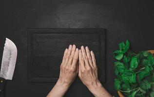 Green fresh mint leaves on a wooden board and two female hands on a black wooden table, top view photo