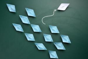 a group of blue paper boats are sailing in one direction, one white one is sailing in the opposite direction. photo