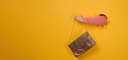 female hand is holding a small golden clutch bag with a metal chain on a yellow background. Part of the body sticking out of a torn hole in a paper background photo