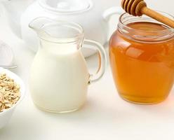 leche fresca en un decantador de vidrio transparente y un tarro de miel sobre una mesa blanca, un producto saludable, desayuno foto