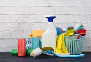 sponges, plastic brushes and bottles of detergents on a blue wooden table. Household cleaning items on white brick wall background photo