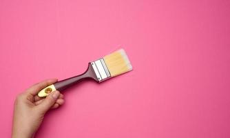 female hand hold new paint brush with wooden handle on a pink background, close up photo
