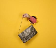 female hand is holding a small golden clutch bag with a metal chain on a yellow background. Part of the body sticking out of a torn hole in a paper background photo