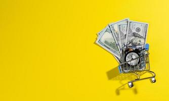 billetes de papel de cien dólares estadounidenses en un carro de metal en miniatura y un despertador negro sobre un fondo amarillo, vista superior foto