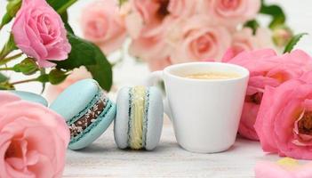 white ceramic cup with coffee and  macarons on a white table, behind a bouquet of pink roses photo