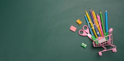 miniature shopping cart full of school office supplies on a green chalk board. Preparing for school, buying pens and notebooks, top view photo