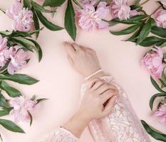 two hands of a young girl with smooth skin and a bouquet of pink peonies on a peach background, top view photo