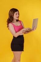 Portrait of Happy young woman holding laptop in her hands while standing isolated over yellow background. Technology and Business concept. photo