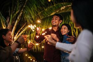 Portrait of Happy Asian group of friends having fun with sparklers outdoor - Young people having fun with fireworks at night time  - People, food, drink lifestyle, new year celebration concept. photo