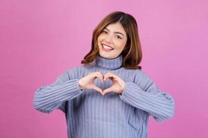 Portrait of Smiling young woman feels happy and romantic shapes heart gesture isolated over pink background